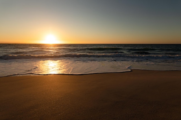 Vista de uma praia de areia e mar calmo com ondas e céu azul ao pôr do sol