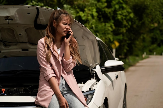 Vista deprimida de una mujer joven sentada en la carretera cerca de un auto roto y hablando con un mecánico por teléfono