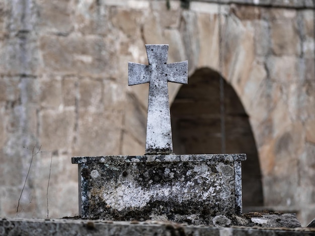 Vista de detalle de una cruz de piedra sobre pedestal del mismo material pero deteriorada por el paso del tiempo encima de un sepulcro en la puerta de una iglesia