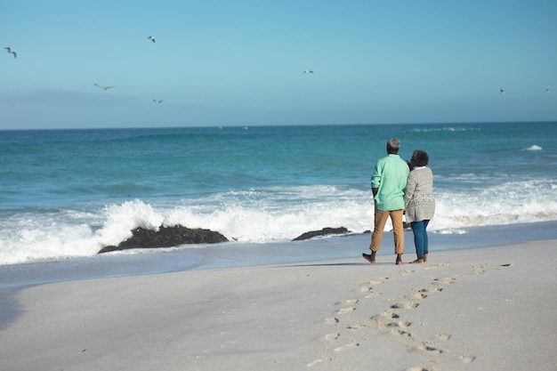 Vista distante de trás de um casal afro-americano sênior de pé na praia com o céu azul e o mar ao fundo, de mãos dadas, olhando para o horizonte