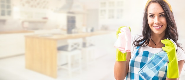 Vista do banner de uma linda mulher sorridente no avental, segurando o detergente e o pano de limpeza nas mãos com luvas de borracha no terço direito da imagem, com cozinha ao fundo.