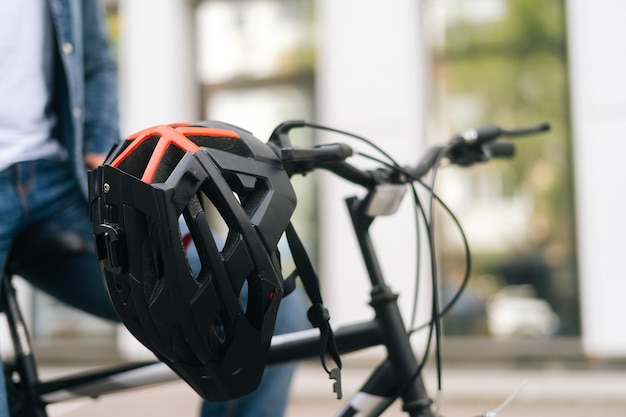 Vista do close-up do capacete da bicicleta pendurado no guidão da bicicleta em pé na rua da cidade num dia de verão em fundo desfocado de edifício urbano. Ciclista irreconhecível sentado no assento da bicicleta.