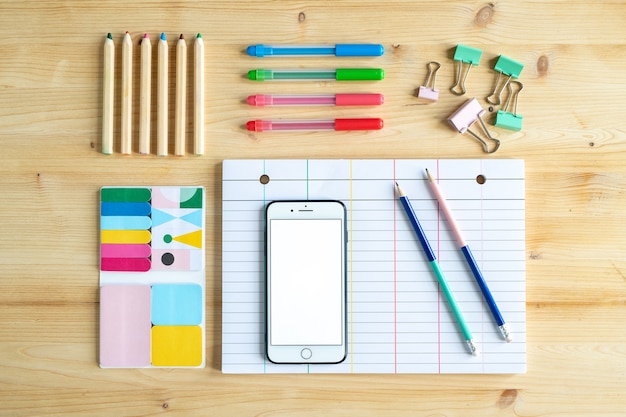 Vista do escritório ou material educacional na mesa de madeira - vários conjuntos de giz de cera, smartphone, clipes e papel pautado