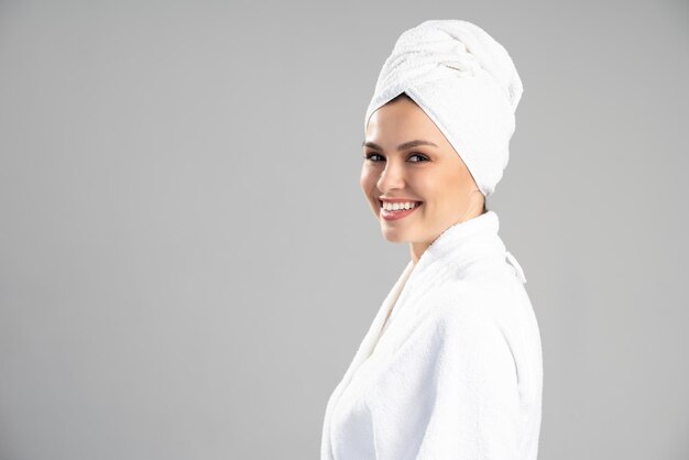 Vista do retrato de uma mulher sorridente, vestindo um roupão branco, posando com uma toalha na cabeça, após tomar banho, isolada sobre o fundo cinza do estúdio