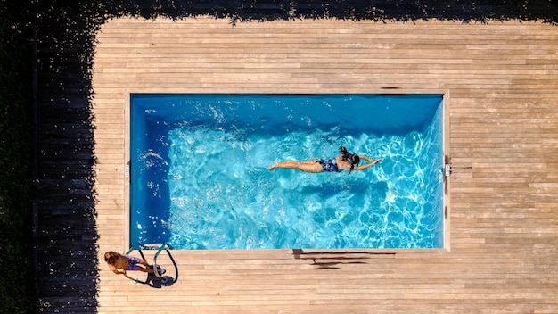 Vista de drone de un niño anónimo de pie en una plataforma de madera cerca de una joven madre nadando en la piscina en un día soleado durante las vacaciones de verano