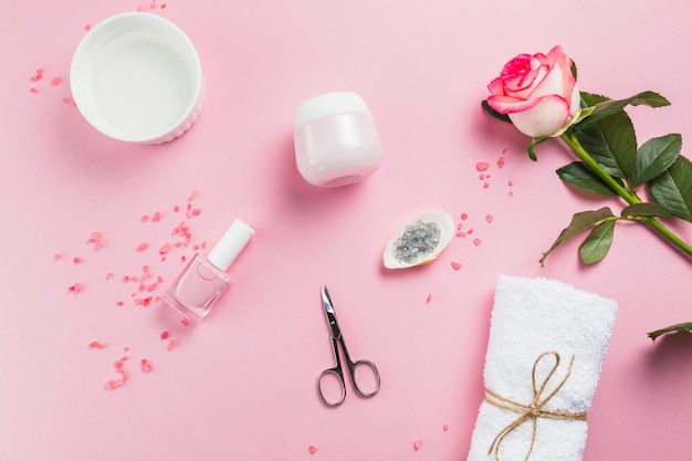 Foto vista elevada do verniz das unhas; tesouras; sal; toalha; flores e creme hidratante na superfície rosa