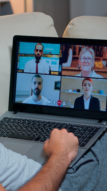 Vista por encima del hombro del hombre en pijama que tiene una conversación en línea con sus compañeros de equipo que trabajan en un proyecto de tecnología en línea usando una computadora portátil, comunicación por Internet por videoconferencia