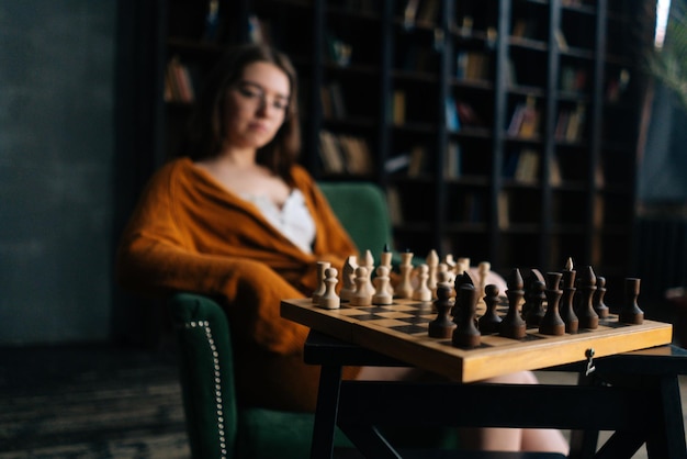 Vista de enfoque selectivo lateral de una joven pensativa con anteojos elegantes pensando en el movimiento de ajedrez sentado en un sillón en una sala de biblioteca oscura