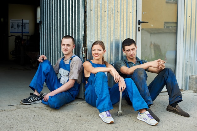 Foto vista de un equipo de mecánicos sentados cerca del taller de reparación. niña sostiene una llave inglesa en su mano, ella sentada entre dos chicos