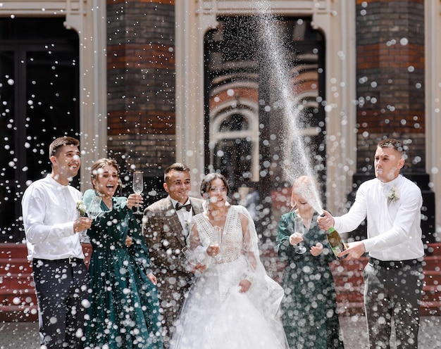 Vista frontal alegre recém-casados com amigos juntos e segurando taças de cristal rindo e olhando para o menino que abre a garrafa e faz respingos de champanhe durante o casamento ao ar livre