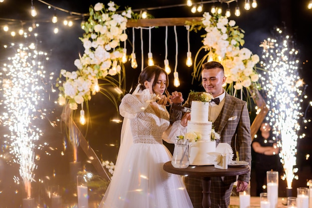 Foto vista frontal de alegres recién casados en elegantes trajes de festival cortando un trozo de pastel y parados en el fondo de un increíble arco de madera