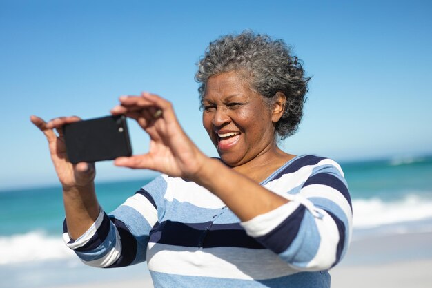 Vista frontal de una anciana afroamericana de pie en la playa con el cielo azul en el fondo, riendo y tomando selfies con un teléfono inteligente