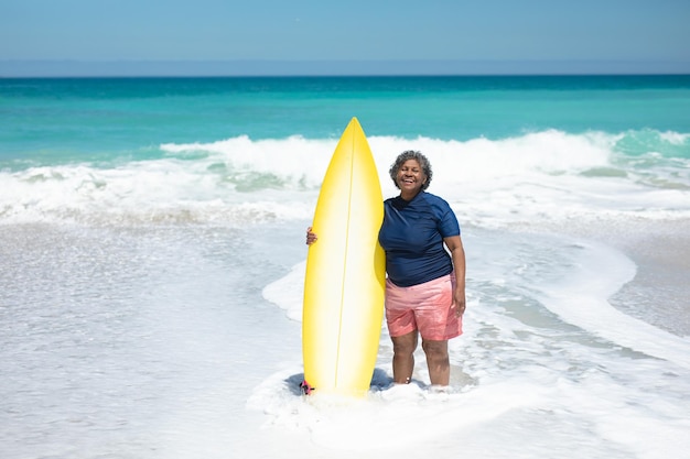 Vista frontal de una anciana afroamericana en una playa al sol, de pie en el mar sosteniendo una tabla de surf, mirando a la cámara y sonriendo, con el cielo azul y el mar tranquilo en el fondo