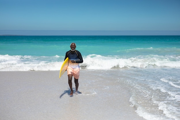 Vista frontal de un anciano afroamericano en una playa al sol, llevando una tabla de surf bajo el brazo, mirando a la cámara y sonriendo, con el cielo azul y el mar en el fondo