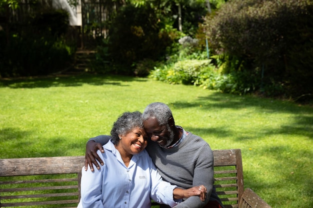 Vista frontal en ángulo alto de una pareja afroamericana mayor en el jardín, sentada en un banco, abrazándose y hablando. Familia disfrutando del tiempo en casa, concepto de estilo de vida