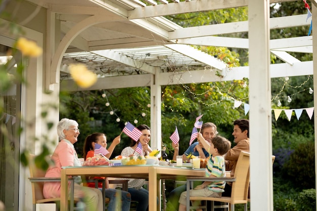 Vista frontal en ángulo bajo de una familia caucásica de varias generaciones sentada afuera en una mesa para cenar, celebrando y ondeando banderas estadounidenses. Familia disfrutando del tiempo en casa, concepto de estilo de vida