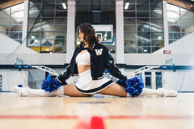 Vista frontal de una animadora morena sosteniendo pompones azules brillantes y haciendo una división en baloncesto