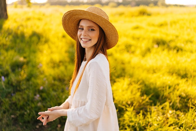 Vista frontal de una atractiva joven feliz con sombrero de paja y vestido blanco de pie posando en un hermoso prado de hierba verde mirando a la cámara