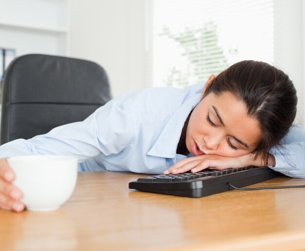 Vista frontal de una bella mujer durmiendo en un teclado mientras sostiene una taza de café