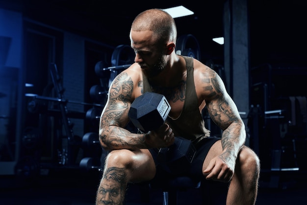 Vista frontal del bíceps de entrenamiento culturista sin camisa con mancuernas en un banco. Cerca de deportista musculoso con cuerpo perfecto posando en el gimnasio en un ambiente oscuro. Concepto de culturismo.