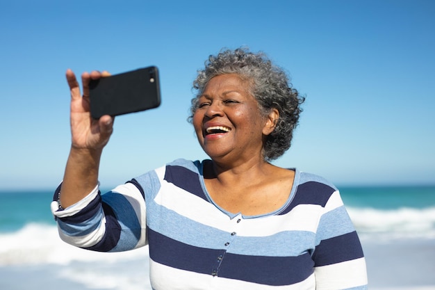 Vista frontal de cerca de una anciana afroamericana de pie en la playa con el cielo azul en el fondo, riendo y tomando una selfie con un teléfono inteligente