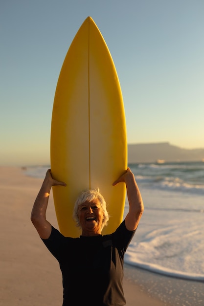 Vista frontal de cerca de una anciana caucásica en la playa al atardecer, de pie en la arena sosteniendo una tabla de surf en la cabeza, mirando a la cámara y riendo