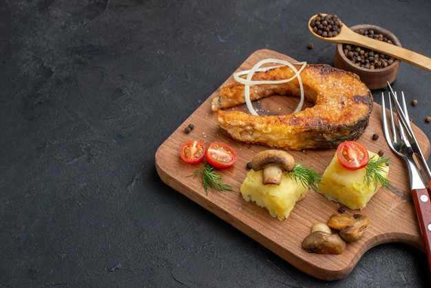 Vista frontal cercana de delicioso pescado frito y champiñones tomates verdes sobre tabla de cortar de madera cubiertos pimienta sobre superficie negra