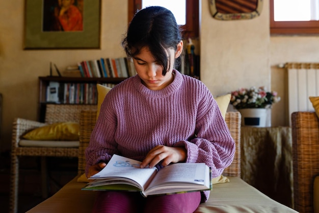 Foto vista frontal de una chica concentrada en un suéter lila leyendo un libro en un sillón amarillo