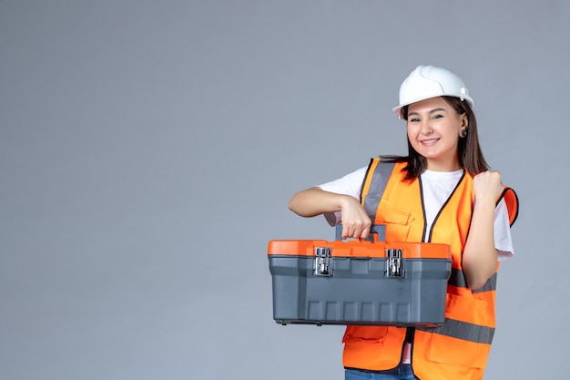 Vista frontal del constructor femenino con caja de herramientas pesada en pared gris