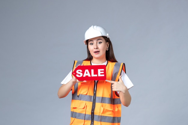 Vista frontal del constructor femenino que sostiene el tablero de venta rojo en la pared blanca