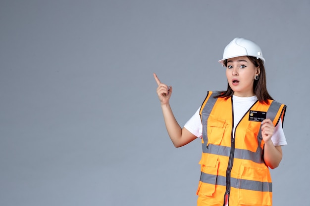 Vista frontal del constructor femenino con tarjeta bancaria negra en sus manos en la pared gris