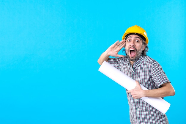 Vista frontal del constructor masculino en casco amarillo con plan sobre fondo azul ingeniero de trabajo de construcción de arquitectura de propiedad de trabajador