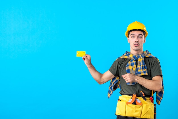 Vista frontal del constructor masculino en casco amarillo sosteniendo una tarjeta bancaria sobre fondo azul dinero arquitectura edificios trabajador constructor color plano
