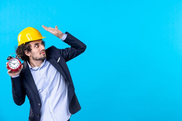 Vista frontal constructor masculino en casco y traje sosteniendo relojes en el fondo azul trabajo de diseño de negocios constructor de arquitectura tiempo de construcción trabajo