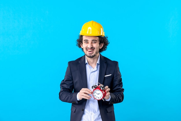 Vista frontal del constructor masculino en casco y traje sosteniendo relojes sobre fondo azul constructor edificio arquitectura trabajo diseño empresarial