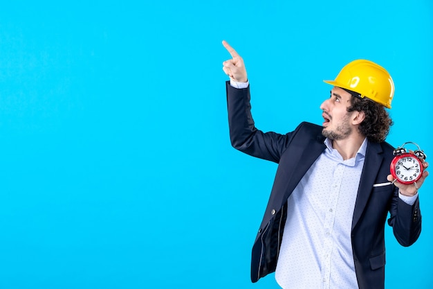 Vista frontal del constructor masculino en casco y traje sosteniendo relojes sobre fondo azul trabajo arquitectura trabajo diseño empresarial constructor edificios tiempo