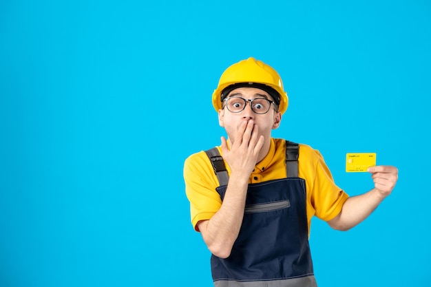 Vista frontal del constructor masculino sorprendido en uniforme y casco con tarjeta de crédito en azul