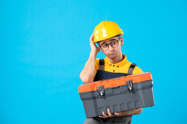 Vista frontal del constructor masculino en uniforme con caja de herramientas en la pared azul