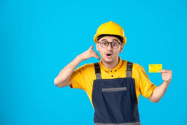 Vista frontal del constructor masculino en uniforme con tarjeta de crédito amarilla en sus manos en la pared azul