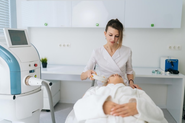 Vista frontal de una cosmetóloga realizando un procedimiento de fotorejuvenecimiento para mujeres en una clínica de belleza Concepto de hardware tecnológico para cambios anti-envejecimiento de la piel de la cara y el cuello