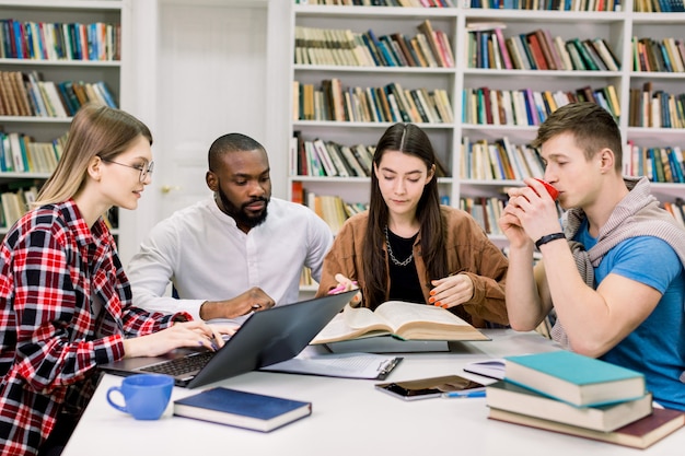 Vista frontal de cuatro estudiantes modernos y elegantes de raza mixta que se sientan en la sala de lectura de la biblioteca y se preparan para exámenes o proyectos científicos.