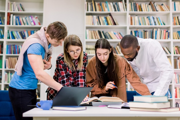 Foto vista frontal de cuatro jóvenes multiétnicos concentrados, estudiantes universitarios que se preparan para su tarea en la sala de la biblioteca