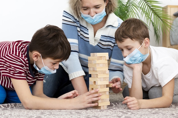 Vista frontal da mãe tocando jenga com crianças em casa enquanto usava máscaras médicas