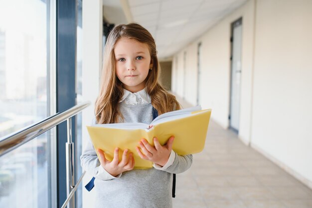 Vista frontal da menina da escola loira bonita segurando muitas notas coloridas e livros Menina adolescente inteligente sorrindo para a câmera em pé no corredor da escola internacional