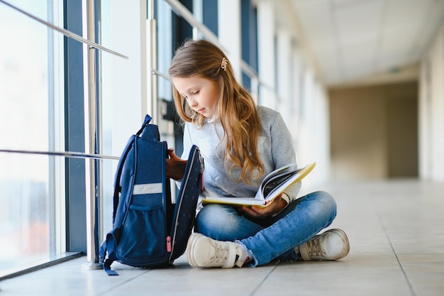 Vista frontal da menina linda escola entre corredor na escola segurando notas nas mãos Menina engraçada e feliz sorrindo para a câmera descansando depois das aulas na escola primária