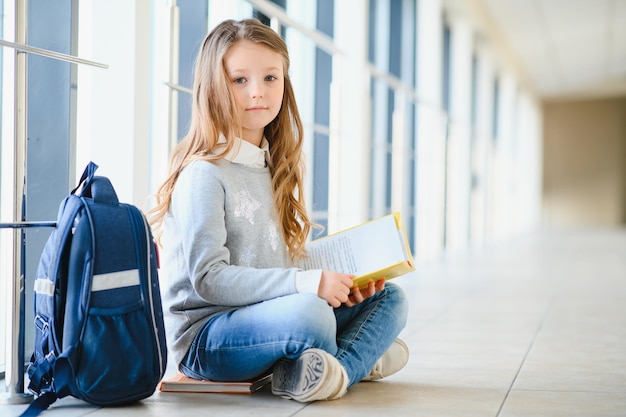 Vista frontal da menina linda escola entre corredor na escola segurando notas nas mãos Menina engraçada e feliz sorrindo para a câmera descansando depois das aulas na escola primária