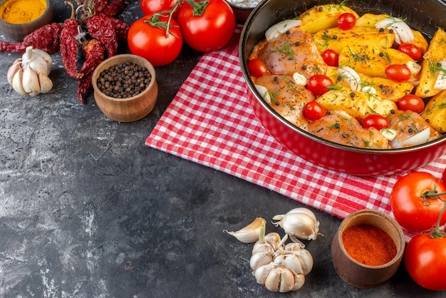 Vista frontal da refeição de frango cru picante com batatas legumes em panela na toalha vermelha despojada e pimentas secas alhos tomates gengibre amarelo em fundo cinza