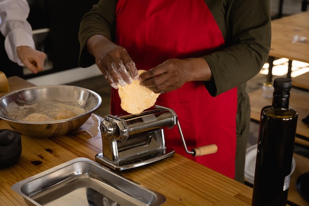 Vista frontal da seção intermediária de uma mulher afro-americana sênior vestindo um avental vermelho preparando comida em uma aula de culinária, colocando massa em uma máquina de macarrão, a chef caucasiana instruindo-a visível
