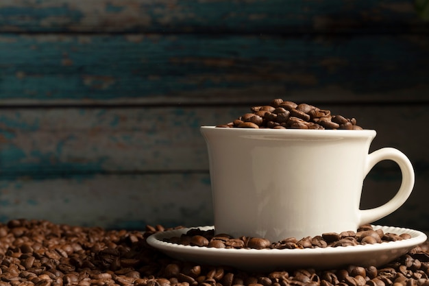 Foto vista frontal da xícara de café cheia de grãos de café em uma mesa com grãos de café espalhados.