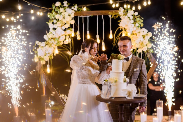 Vista frontal de recém-casados alegres em trajes elegantes de festival cortando um pedaço de bolo e de pé no fundo de um incrível arco de madeira